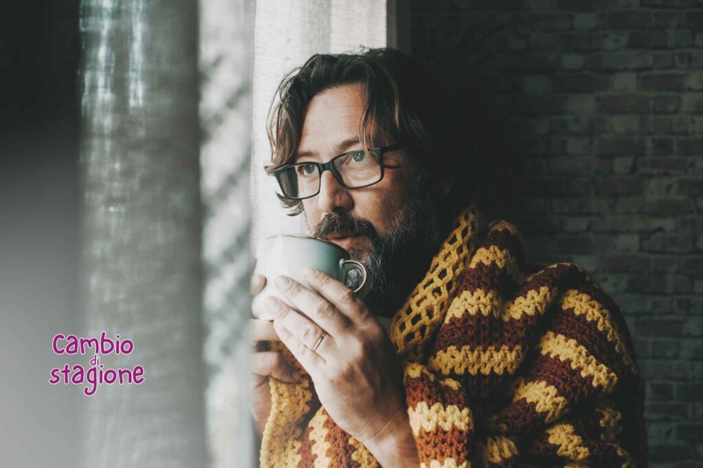 uomo che sente freddo alla finestra beve una bevanda calda